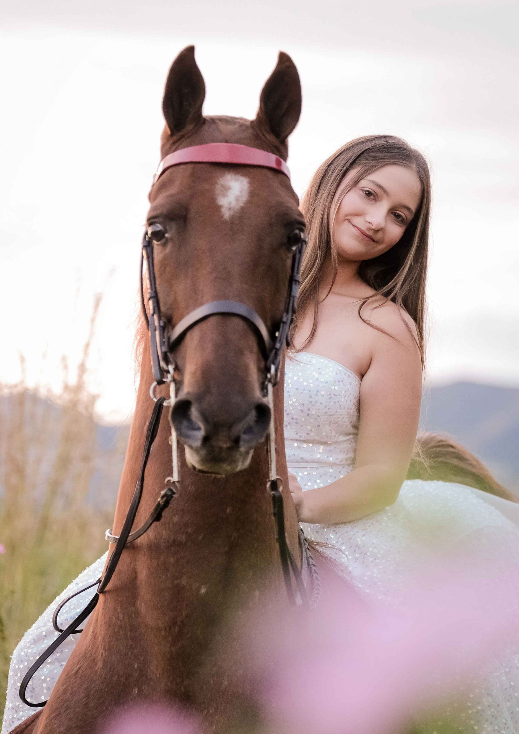 Teenage girl in a sparkly white dress sitting on a chestnut horse among pink flowers with mountains behind