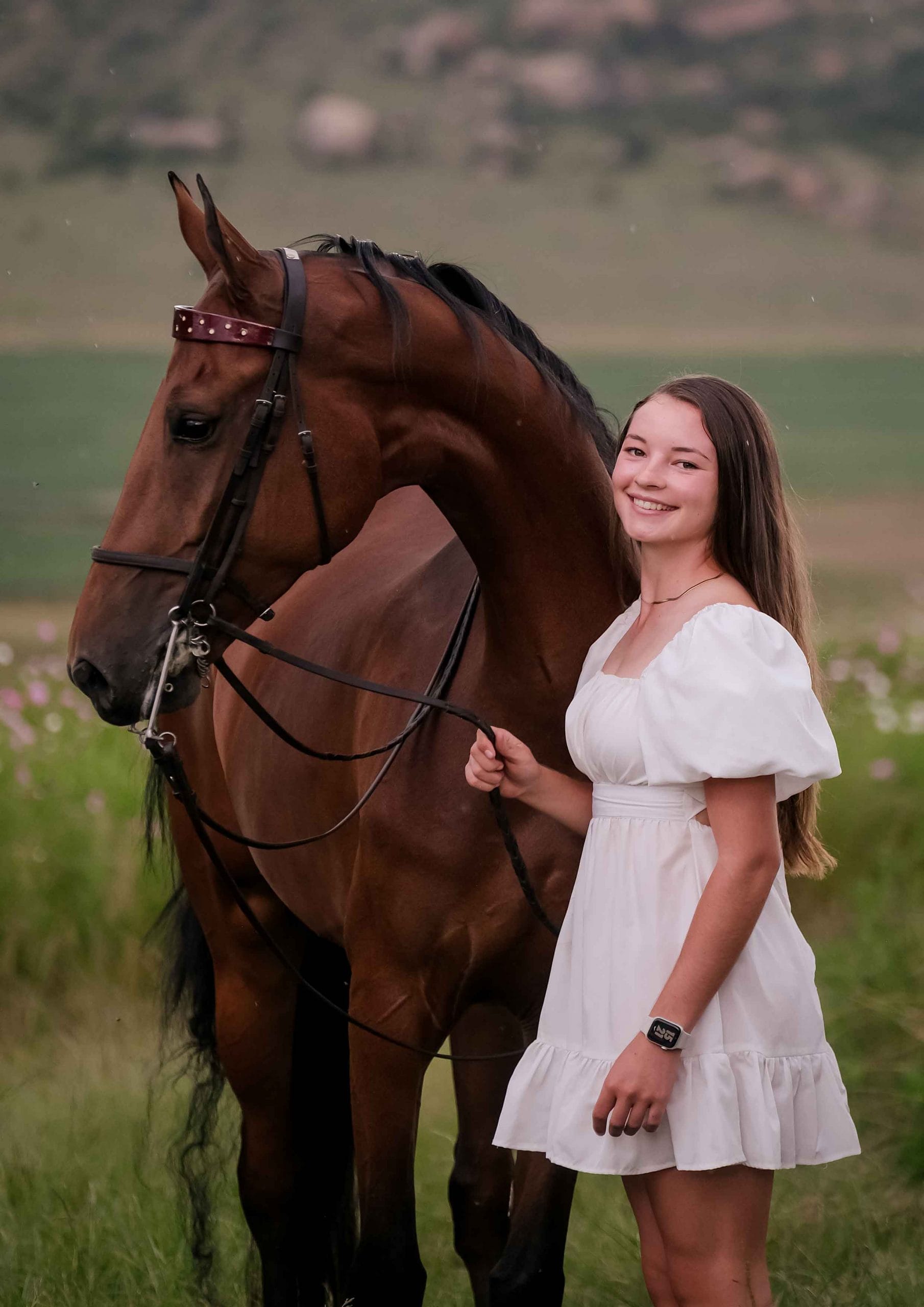Smiling teenage girl in a white puff-sleeve dress standing beside a bay horse in a field of wildflowers
