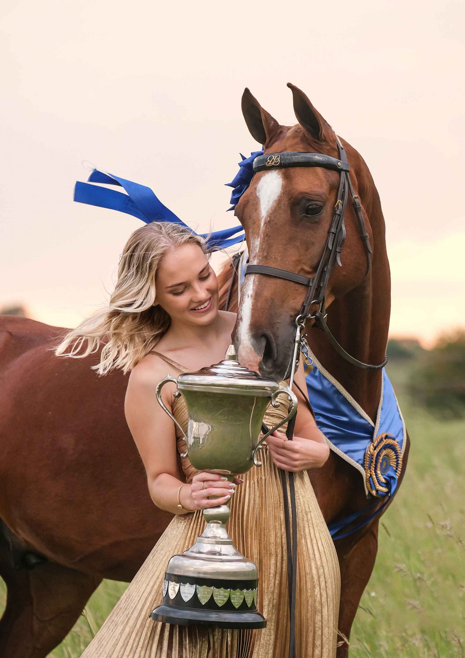 Blonde woman in a gold gown holding a trophy beside a chestnut horse wearing a blue rosette at sunset