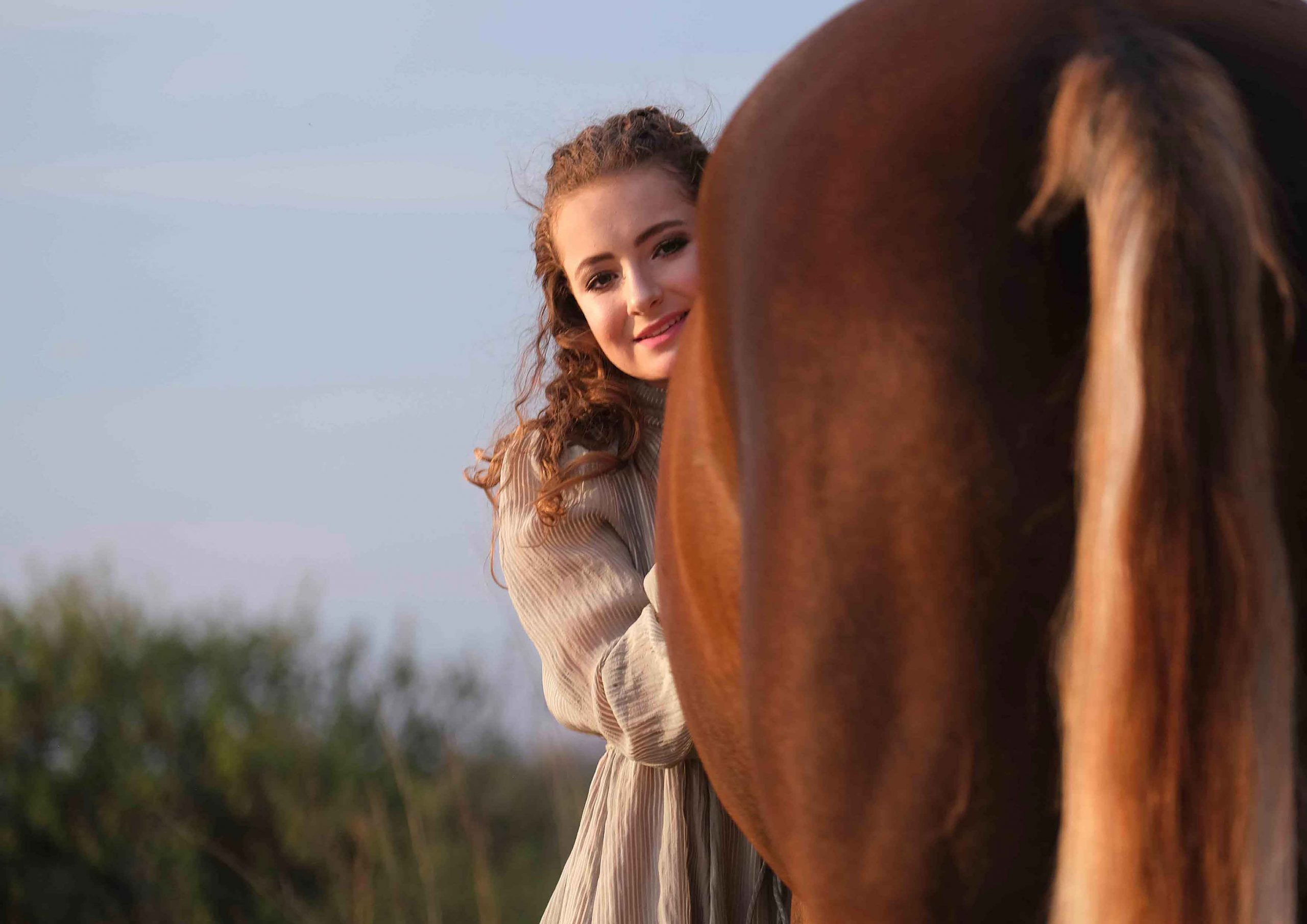 Curly-haired woman peeking out playfully from behind a chestnut horse's flank at sunset