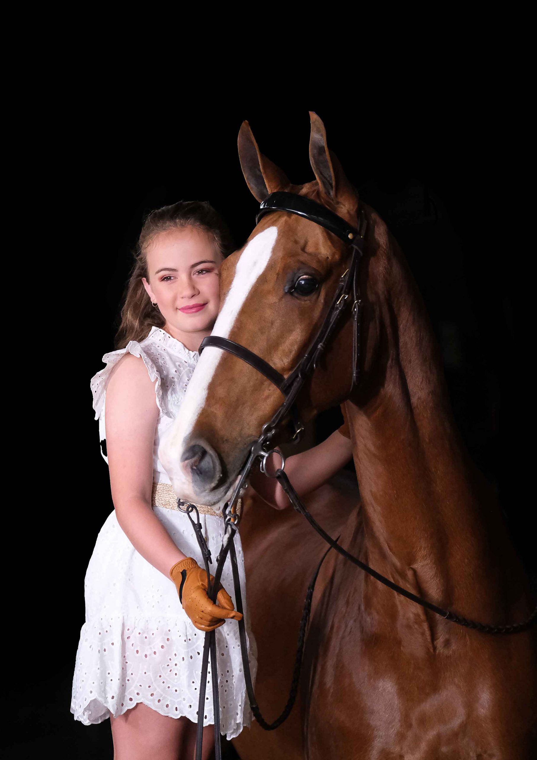 Teenage girl in a white dress leaning against a chestnut horse's face in a dark studio portrait