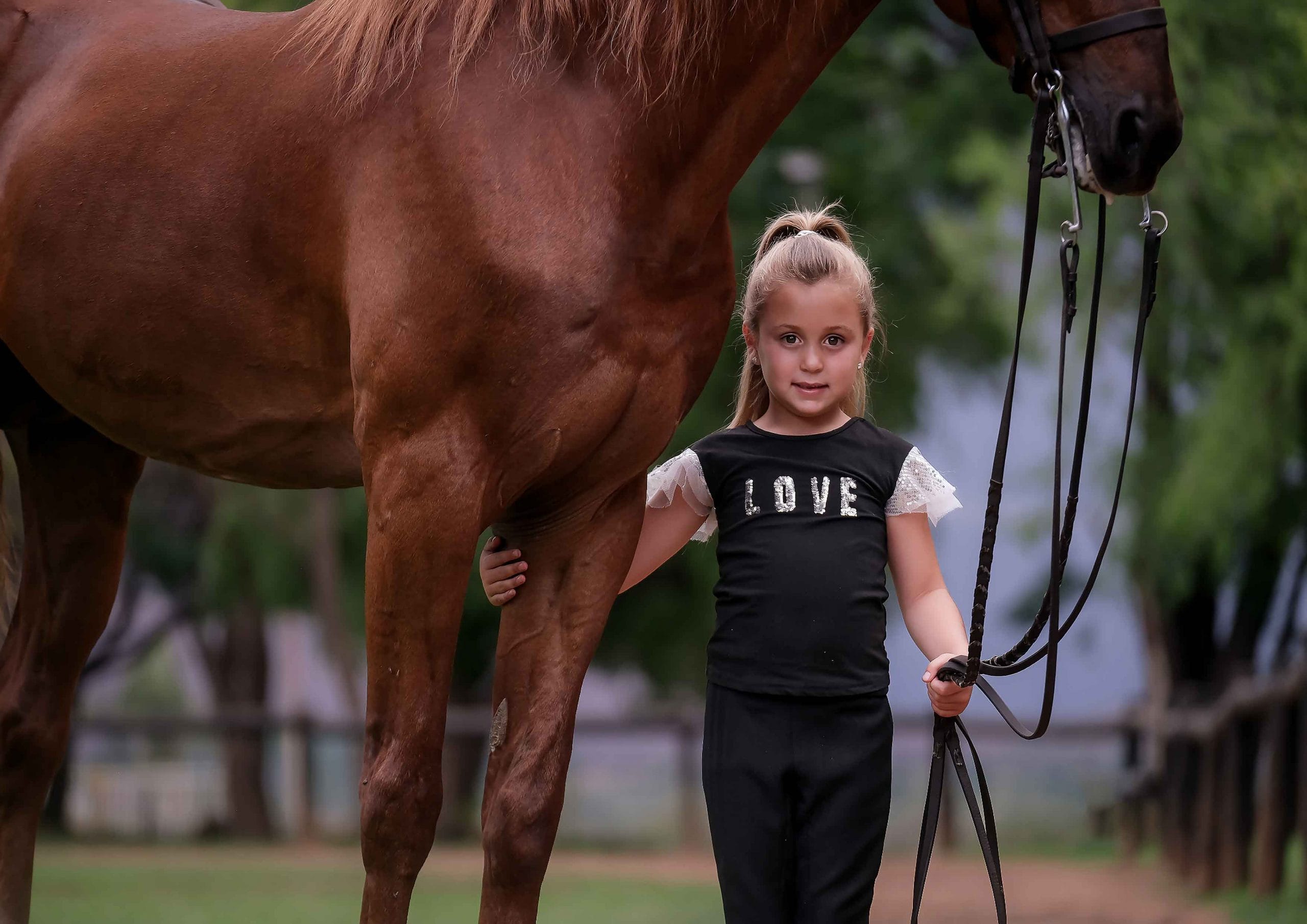 Young girl in a black Love t-shirt standing beside a chestnut horse, holding its reins on a farm