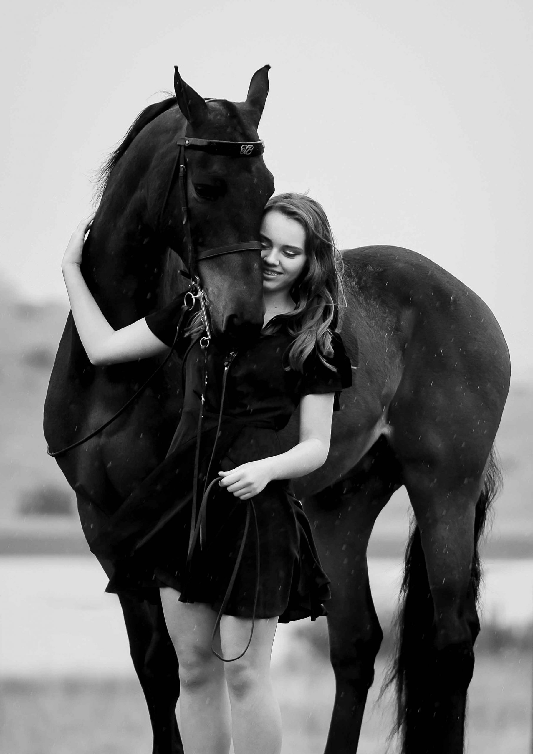 Black and white photo of a smiling teenage girl in a dark dress embracing the head of a dark bay horse