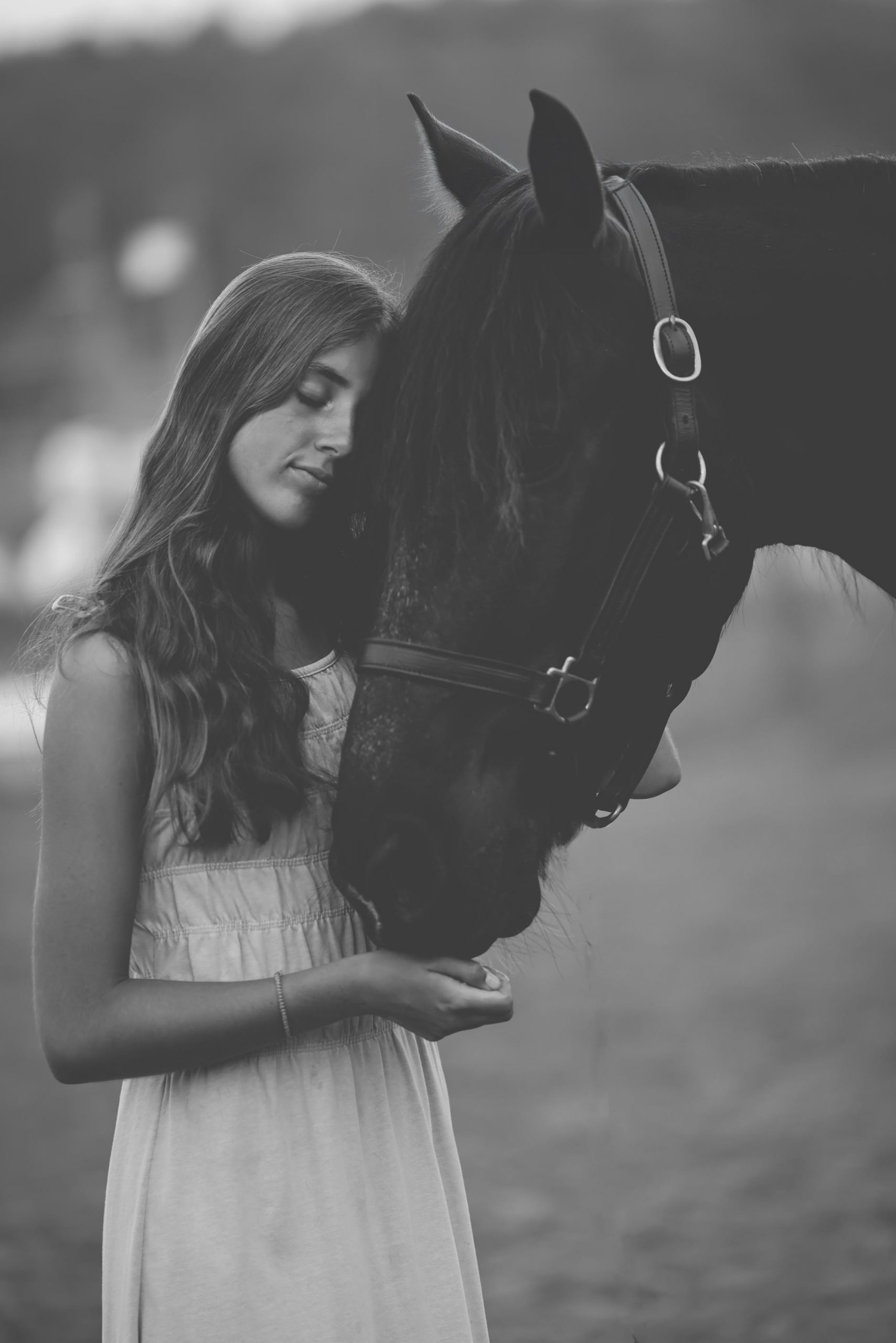 Young woman with wavy hair closing her eyes as she rests against a dark horse's face, black and white portrait
