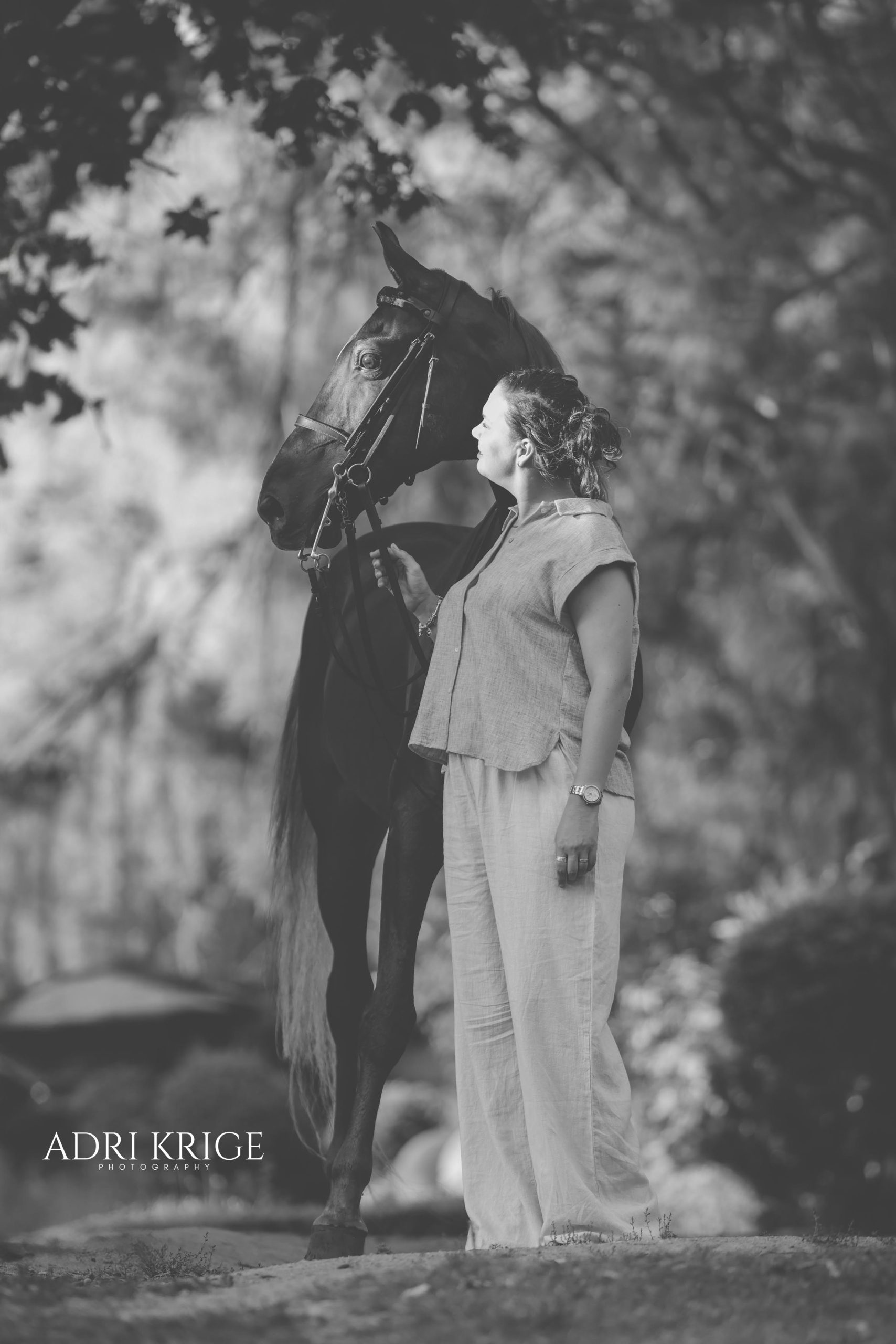 Black and white photo of a woman standing face to face with a dark horse beneath trees in a garden setting
