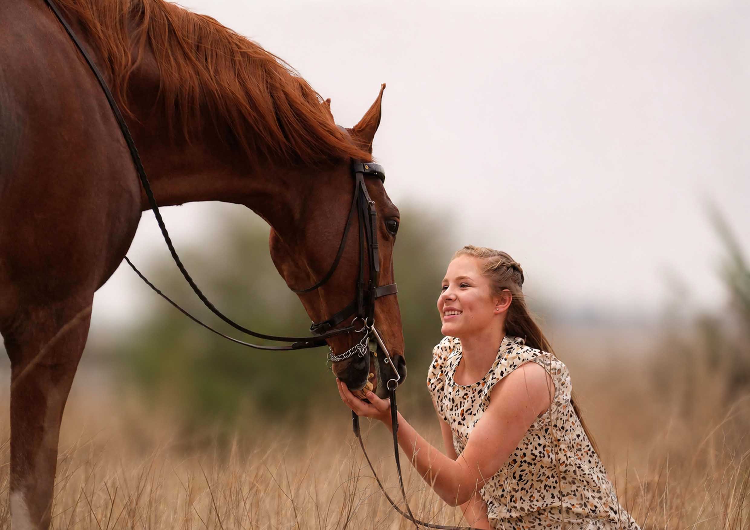 Girl in a patterned dress kneeling and smiling while holding the bridle of a chestnut horse in a golden field