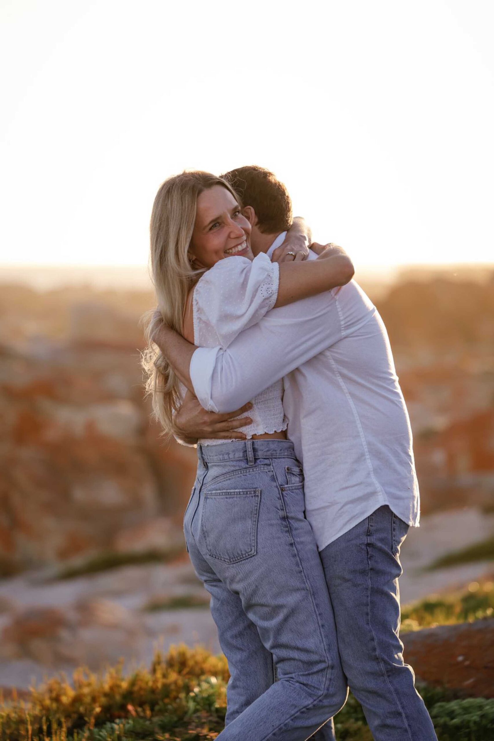 Newly engaged couple in white shirts and jeans hugging at golden hour, the smiling woman showing her engagement ring, engagement photoshoot in Jeffreys Bay by Adri Krige, Eastern Cape wedding photographer