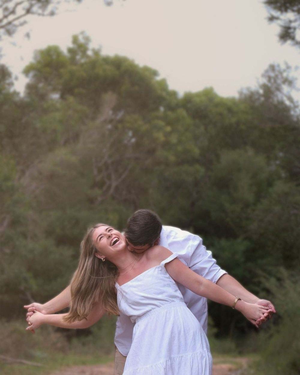 Laughing woman in a white dress holding hands with her partner as he kisses her neck among green trees, anniversary couples photoshoot by Adri Krige