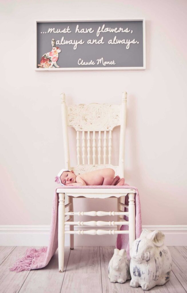Newborn baby in a white headband with a pink flower lying on a pink blanket on a carved vintage white chair in a pastel nursery, with a framed floral quote above and white bunny ornaments beside the chair, photographed by Adri Krige Photography
