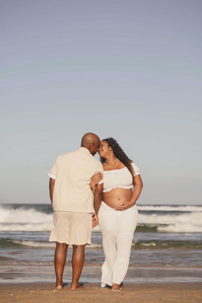 Expectant couple dressed in white and cream standing barefoot at the water’s edge, foreheads touching and smiling, her hand resting on her bare bump, with breaking waves and a wide blue sky, maternity photoshoot by Adri Krige Photography