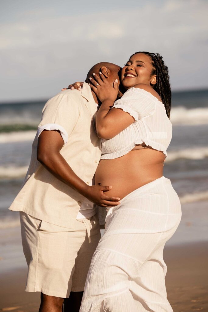 Joyful expectant mother in a white crop top and long skirt hugging her partner close on the beach, eyes closed and smiling, as he cradles her bare bump with the waves behind them, maternity photoshoot by Adri Krige Photography