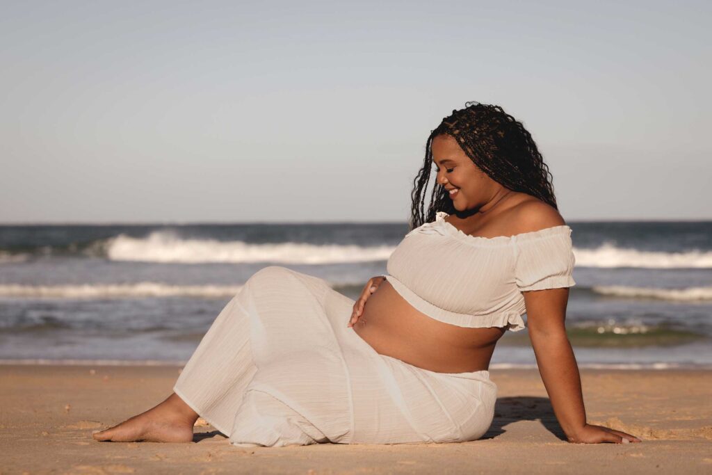 Expectant mother in a white off-the-shoulder crop top and long skirt sitting on the sand by the sea, smiling down at her bare bump as she cradles it, with waves breaking behind her, maternity photoshoot by Adri Krige Photography