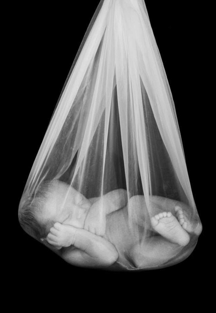 Black and white photo of a sleeping newborn baby curled up inside a soft sheer tulle wrap against a black background, with tiny feet and hands visible through the fabric, photographed by Adri Krige Photography