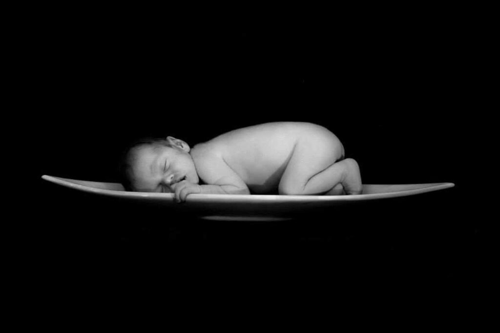 Black and white photo of a sleeping newborn baby curled up on a wide white dish against a black background, with one tiny hand resting on the rim, photographed by Adri Krige Photography