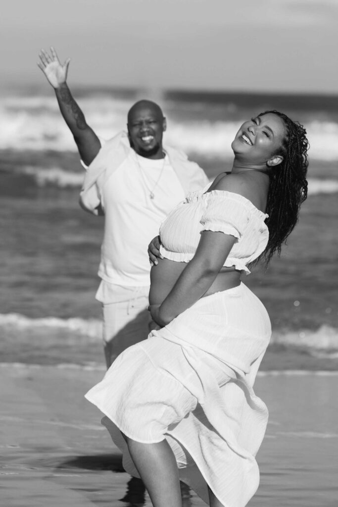 Black and white photo of a laughing expectant mother in a flowing white skirt and crop top holding her bump on the beach while her partner waves and laughs in the background by the waves, maternity photoshoot by Adri Krige Photography