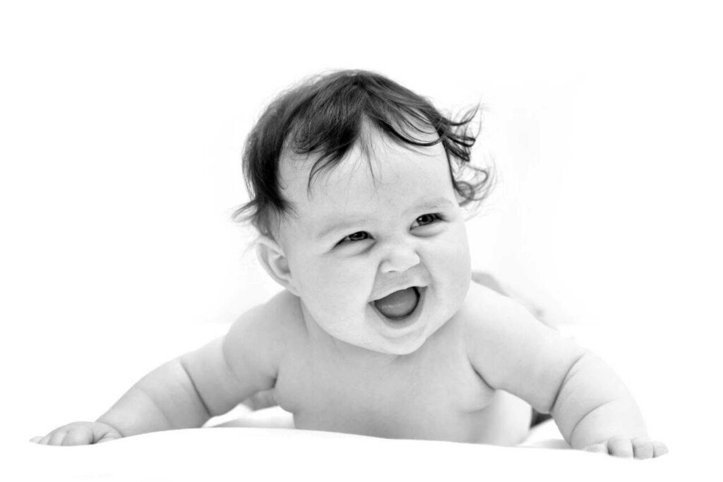 Black and white portrait of a laughing baby with dark curly hair lying on their tummy and pushing up on chubby arms against a white background, photographed by Adri Krige Photography