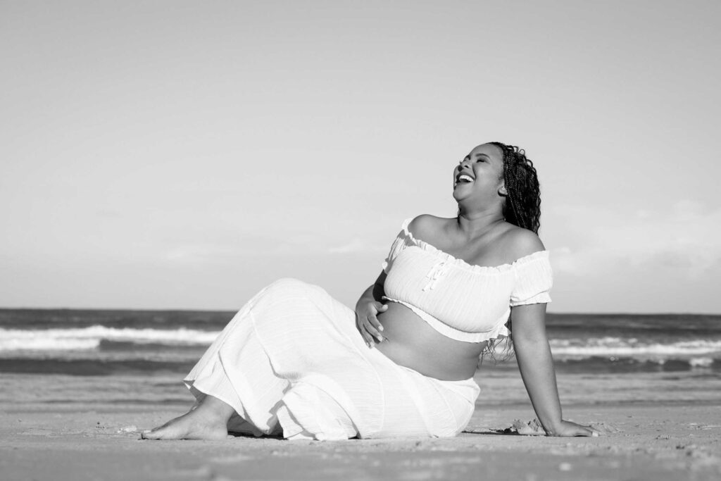Black and white photo of an expectant mother in a white crop top and long skirt sitting on the sand and laughing with her head tilted back, one hand on her bump, with ocean waves behind her, maternity photoshoot by Adri Krige Photography