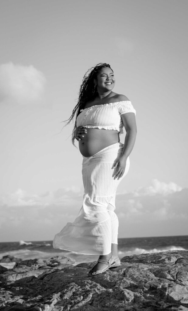 Black and white portrait of a smiling expectant mother in a white crop top and flowing skirt standing on coastal rocks above the sea, one hand on her bump and her long braids blowing in the wind, maternity photoshoot by Adri Krige Photography