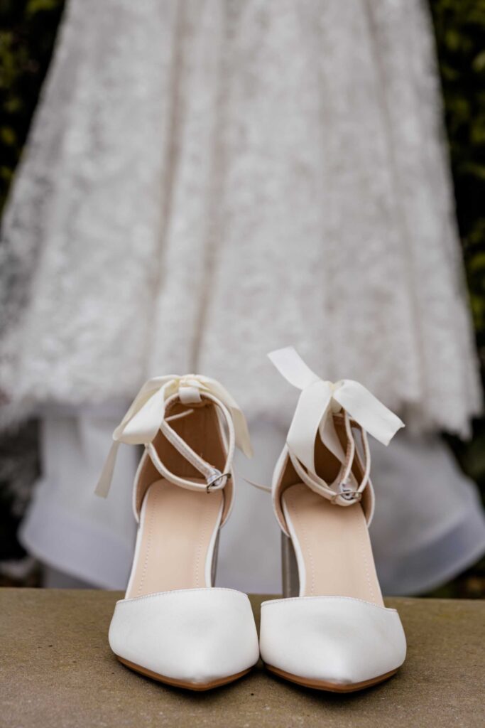 Bridal shoes and lace wedding dress White pointed bridal shoes with satin ribbon bows in front of a hanging lace wedding dress, photographed by Adri Krige, wedding photographer St Francis Bay