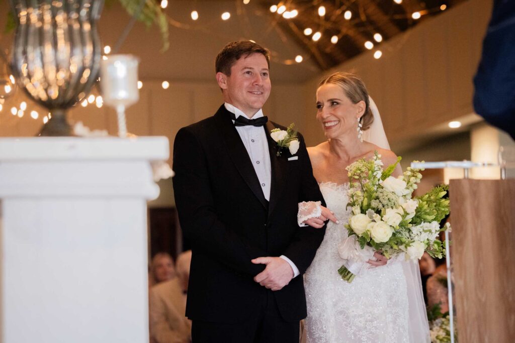Smiling bride with her white bouquet looking up at her groom in a black tuxedo as they stand together at their ceremony under warm fairy lights at St Francis Links, photographed by Adri Krige, wedding photographer