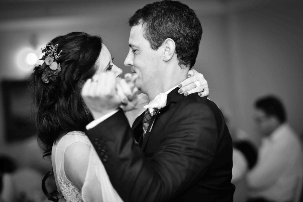 Black and white photo of a bride with flowers in her dark hair and her groom smiling as they hold each other close during their first dance, photographed by Adri Krige, wedding photographer