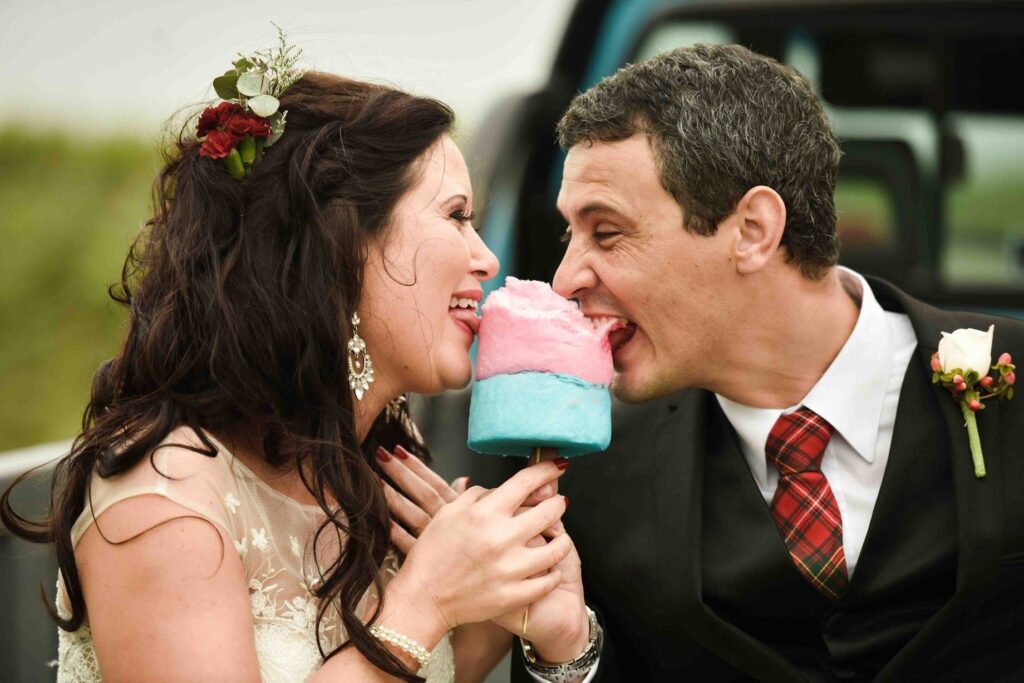 Laughing bride and groom in a red tartan tie playfully biting into the same pink and blue candy floss on their wedding day, photographed by Adri Krige, wedding photographer