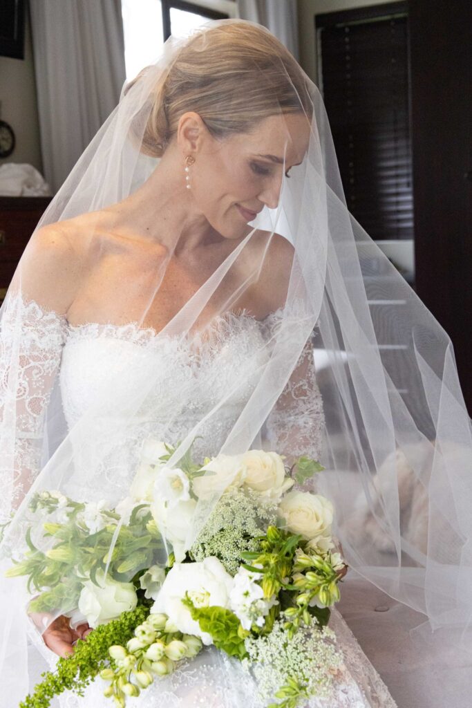 Bride in an off-the-shoulder lace gown sitting beneath her sheer veil, smiling softly as she looks down at her bouquet of white roses, Queen Anne's lace and green blooms, on her wedding day at St Francis Links, photographed by Adri Krige, wedding photographer