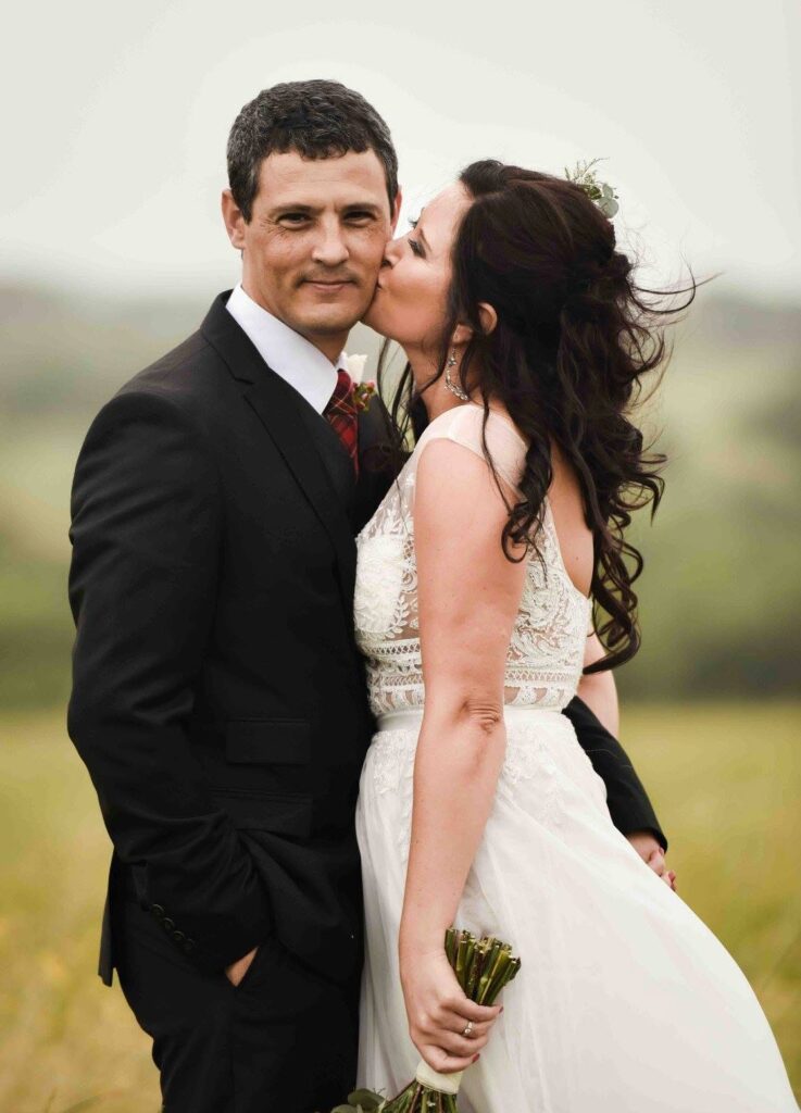 Bride with long dark curls and a lace gown kissing her smiling groom on the cheek as he stands in a dark suit and tartan tie, in a soft green field, photographed by Adri Krige, Garden Route wedding photographer