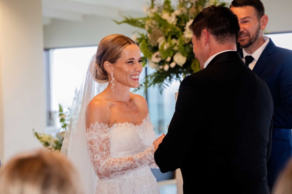 Bride in an off-the-shoulder lace gown and veil beaming at her groom as they hold hands during their wedding vows, with white flowers and greenery behind them, at St Francis Links, photographed by Adri Krige, wedding photographer