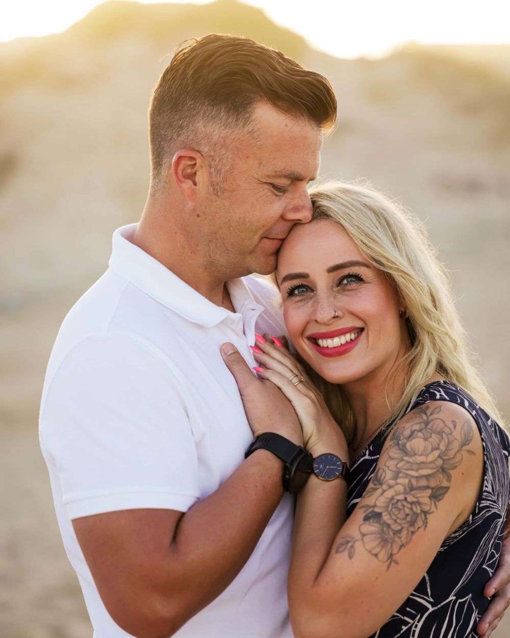 Smiling woman leaning into her partner as he kisses her forehead among sunlit sand dunes, couple photoshoot in Jeffreys Bay by Adri Krige