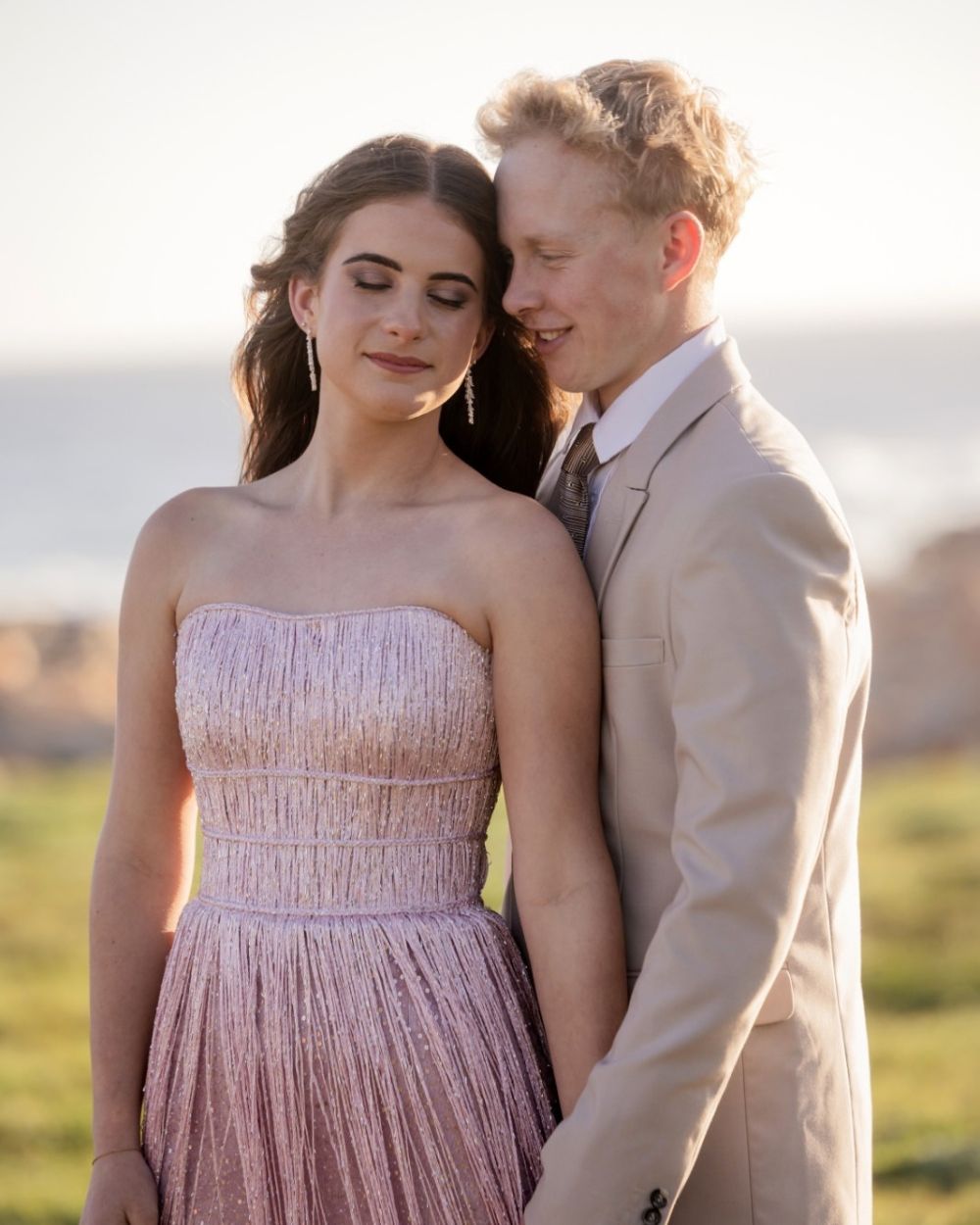 Couple in a blush pink gown and beige suit sharing a tender embrace by the sea at golden hour, couples photoshoot by Adri Krige