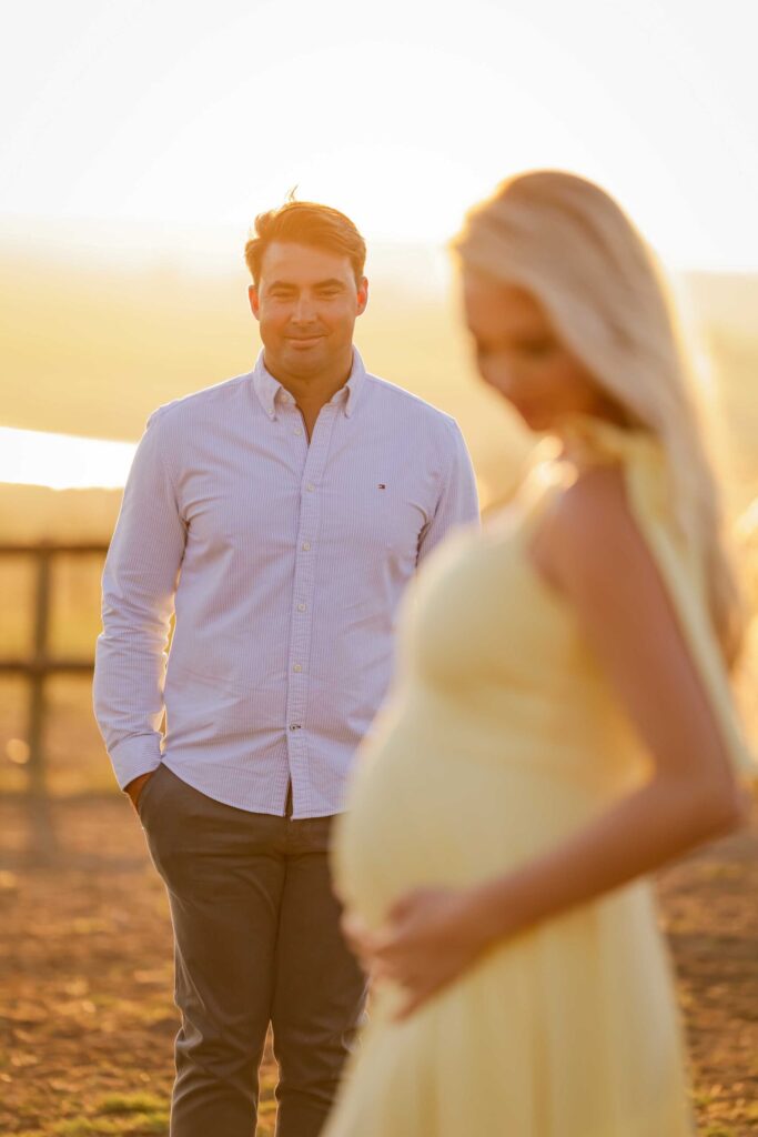 Dad-to-be in a striped shirt smiling softly as he watches his expectant partner, softly out of focus in the foreground, cradle her bump in a yellow dress in hazy golden sunlight, maternity photoshoot by Adri Krige Photography