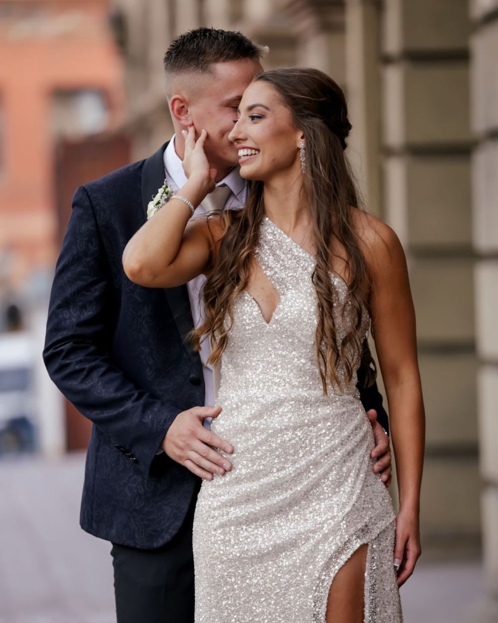 Smiling woman in a sparkling sequin gown touching the face of her partner in a navy suit as he kisses her cheek, engagement session by Adri Krige