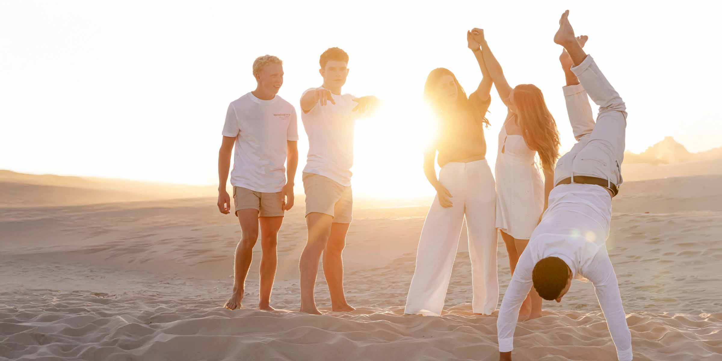 Group of young friends laughing and playing barefoot in the sand dunes at sunset, one doing a handstand, photoshoot in Jeffreys Bay by Adri Krige