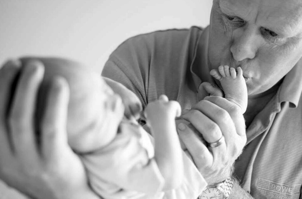 Black and white photo of a father gently kissing the tiny foot of his newborn baby, cradled in his large hands, photographed by Adri Krige Photography