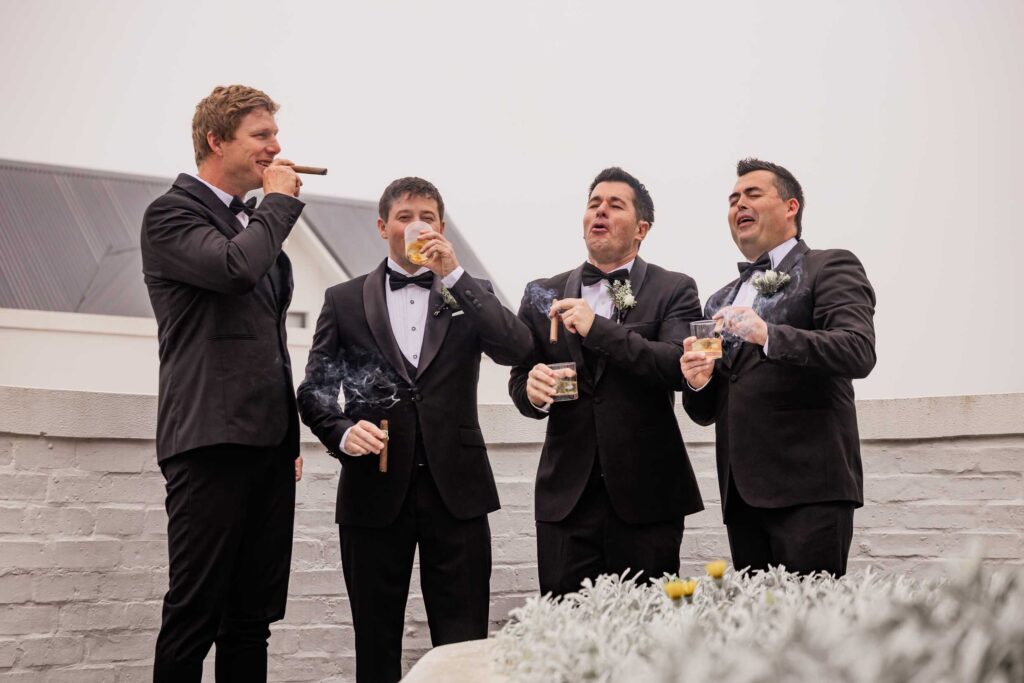 Groom and groomsmen with cigars Groom and three groomsmen in black tuxedos laughing together with cigars and glasses of whisky beside a white brick wall, photographed by Adri Krige, wedding photographer St Francis Bay