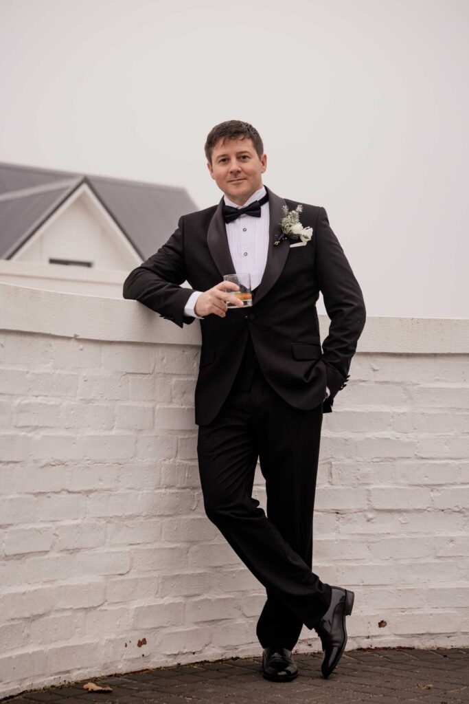 Groom leaning on a white wall Groom in a black tuxedo with a white rose buttonhole leaning on a white brick wall, holding a glass of whisky and smiling, photographed by Adri Krige, wedding photographer St Francis Bay