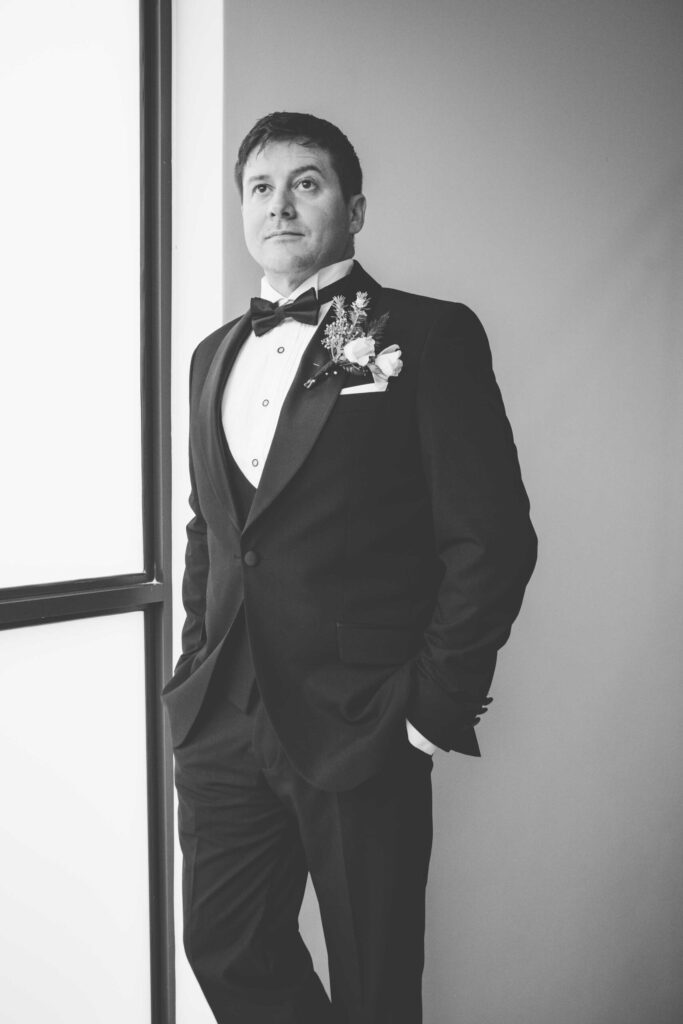Groom portrait by the window Black and white portrait of the groom in a tuxedo and bow tie with a rose buttonhole, standing by a bright window with his hands in his pockets, photographed by Adri Krige, wedding photographer St Francis Bay