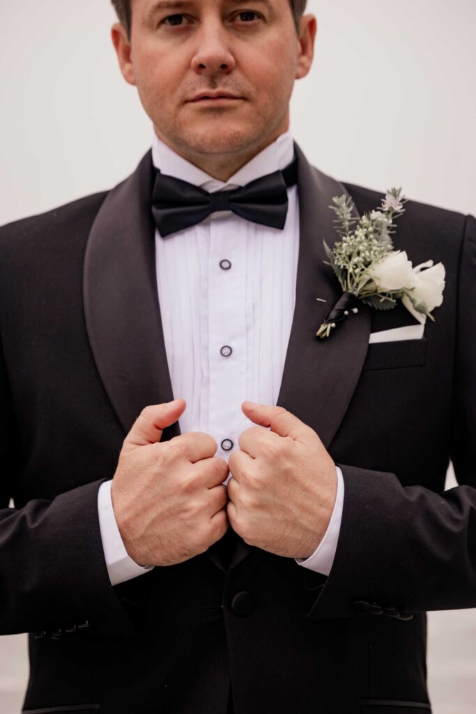 Groom tuxedo and buttonhole detail Close-up of the groom holding the lapels of his black tuxedo, showing his bow tie, pleated shirt and white rose buttonhole, photographed by Adri Krige, wedding photographer St Francis Bay