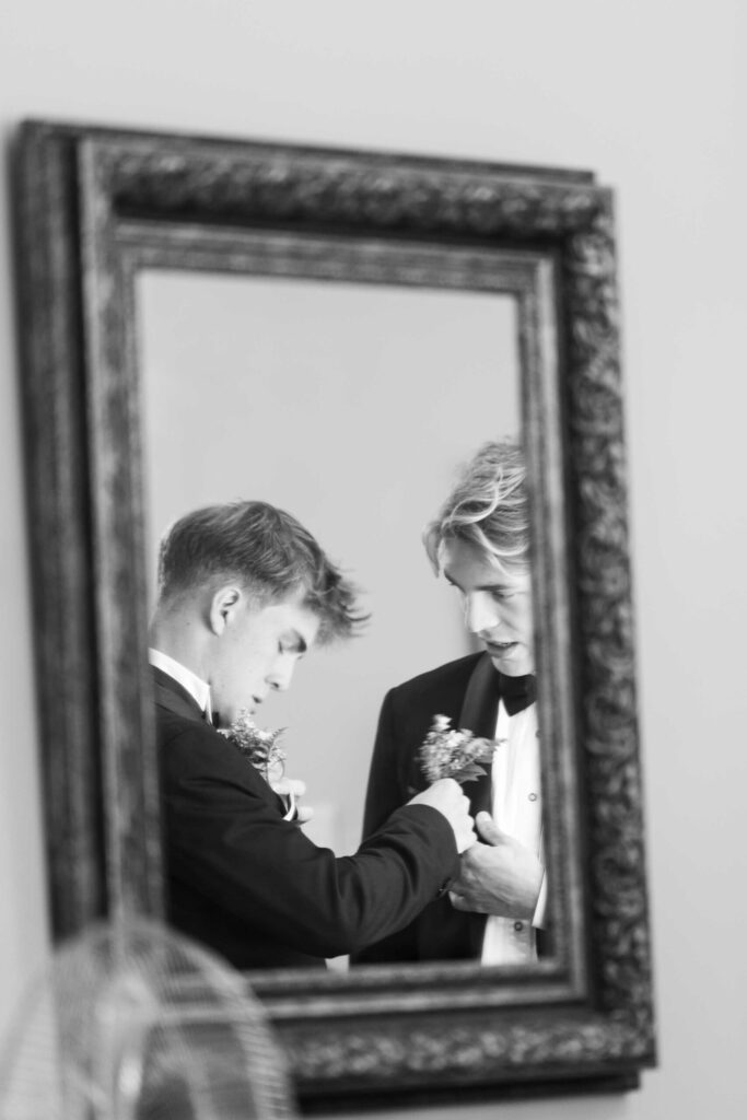Buttonholes in the mirror Black and white photo of two young men in tuxedos reflected in an ornate mirror as they help each other pin on their buttonholes, photographed by Adri Krige, wedding photographer St Francis Bay