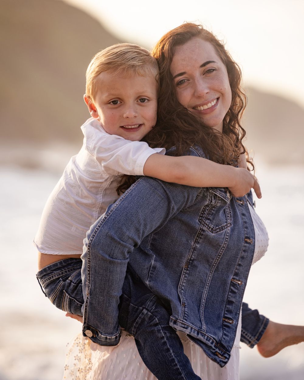 Smiling mother giving her young son a piggyback on the beach at golden hour, holiday family photoshoot by Adri Krige