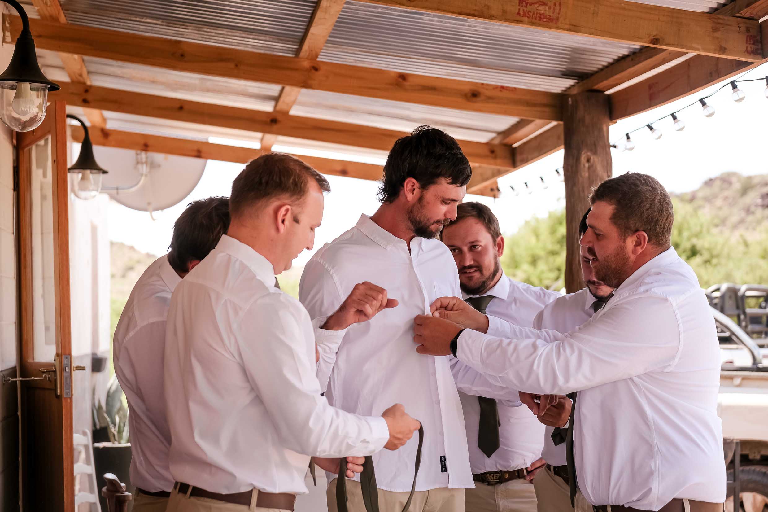Groomsmen in white shirts and olive ties helping the groom button his shirt and tie his tie on a farmhouse stoep, wedding getting-ready photography by Adri Krige, Eastern Cape wedding photographer