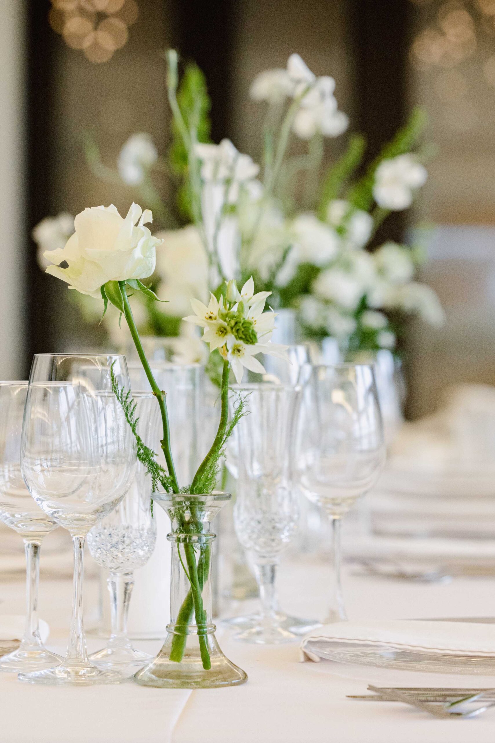A single white rose and white star flowers in a small glass bud vase among crystal wine glasses on a wedding reception table, with white flower arrangements behind, wedding detail photography by Adri Krige, Eastern Cape wedding photographer