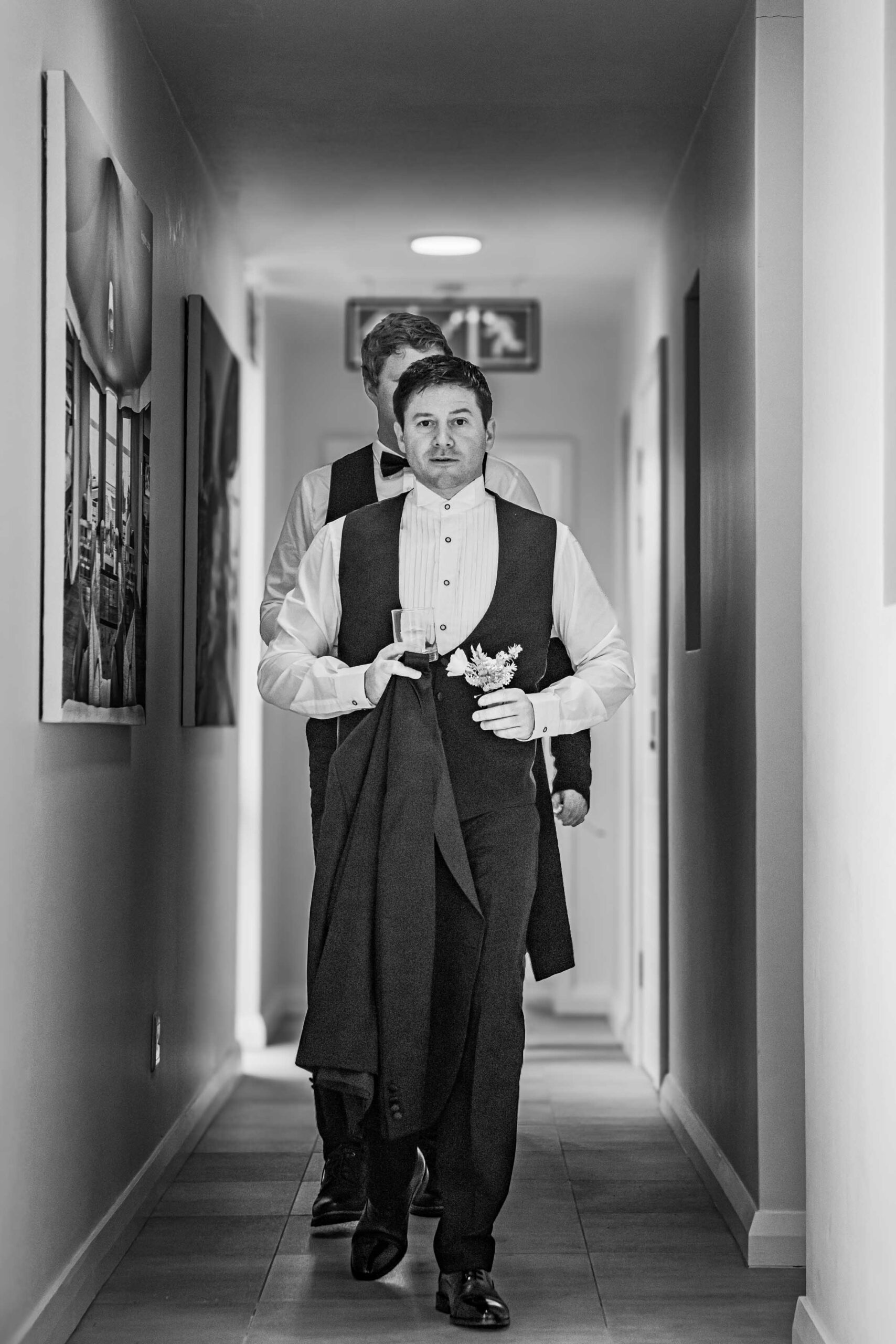 Black and white photo of a groom in a waistcoat and wing-collar shirt walking down a hallway carrying his jacket and buttonhole flowers, followed by a groomsman, wedding photography by Adri Krige, Eastern Cape wedding photographer