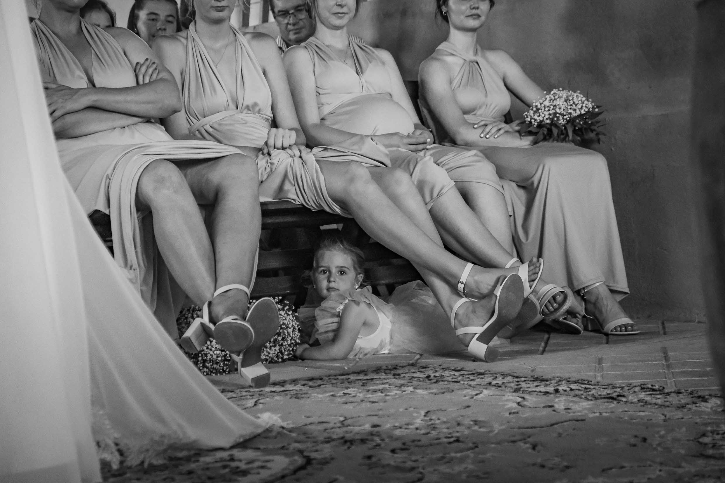 Black and white photo of a little flower girl in a tulle dress lying on the floor beneath the bench of seated bridesmaids during the wedding ceremony, wedding photography by Adri Krige, Eastern Cape wedding photographer