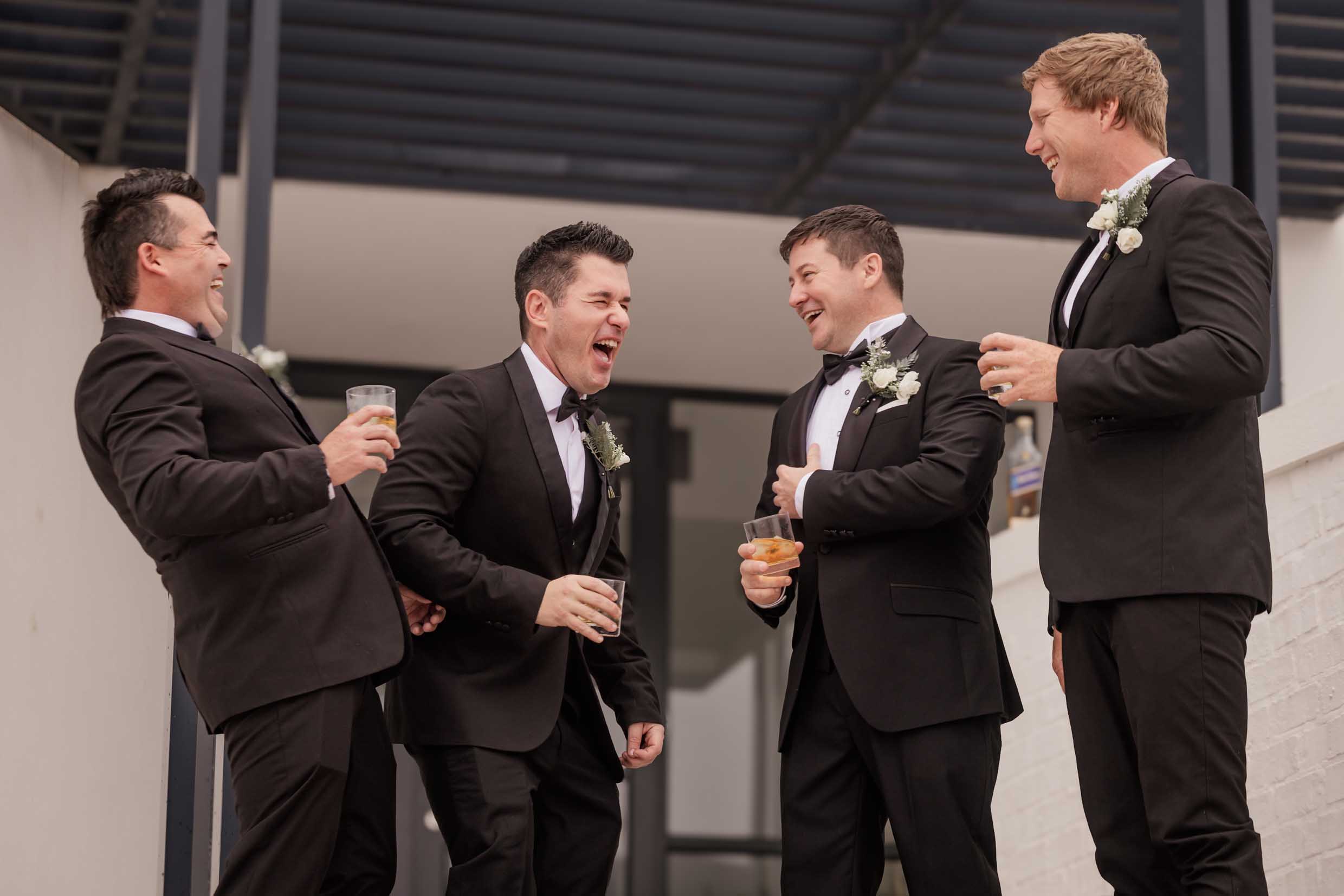 Groom and groomsmen in black tuxedos and white rose buttonholes laughing together with drinks in hand before the wedding, wedding photography by Adri Krige, Eastern Cape wedding photographer
