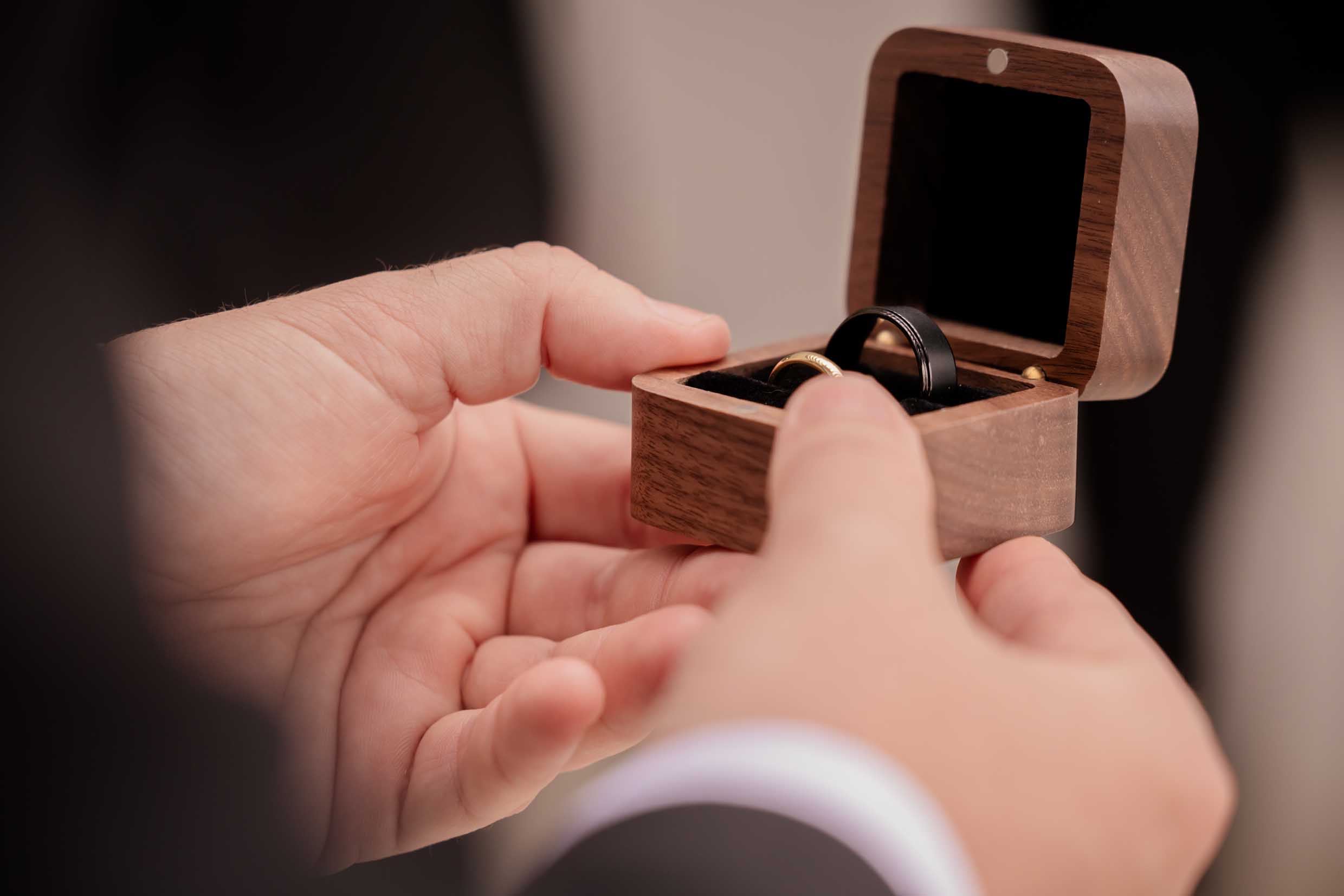 Close-up of hands holding an open walnut ring box with a gold wedding band and a black wedding band inside, wedding detail photography by Adri Krige, Eastern Cape wedding photographer