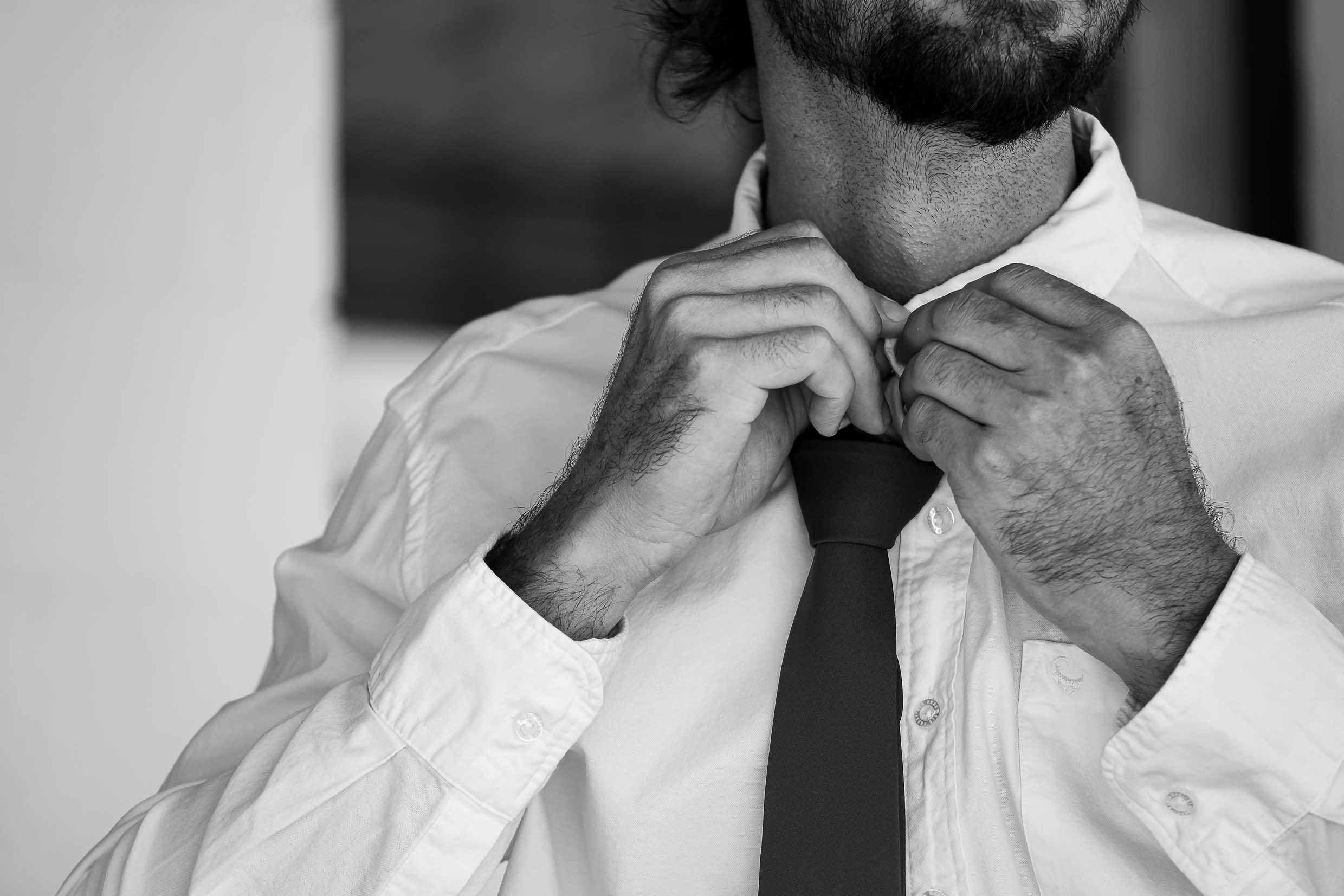 Black and white close-up of a bearded man in a white shirt buttoning his collar over a slim dark tie while getting ready for the wedding, wedding photography by Adri Krige, Eastern Cape wedding photographer
