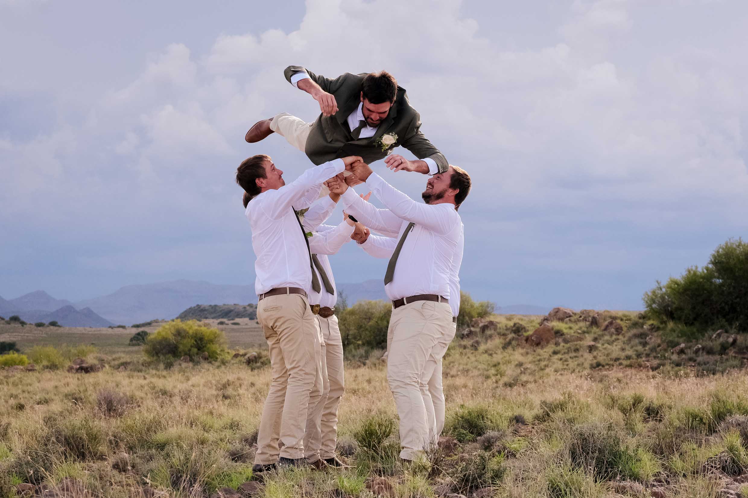 Groomsmen in white shirts and olive ties tossing the groom in an olive jacket high into the air in the open veld, with mountains and storm clouds behind, wedding photography by Adri Krige, Eastern Cape wedding photographer