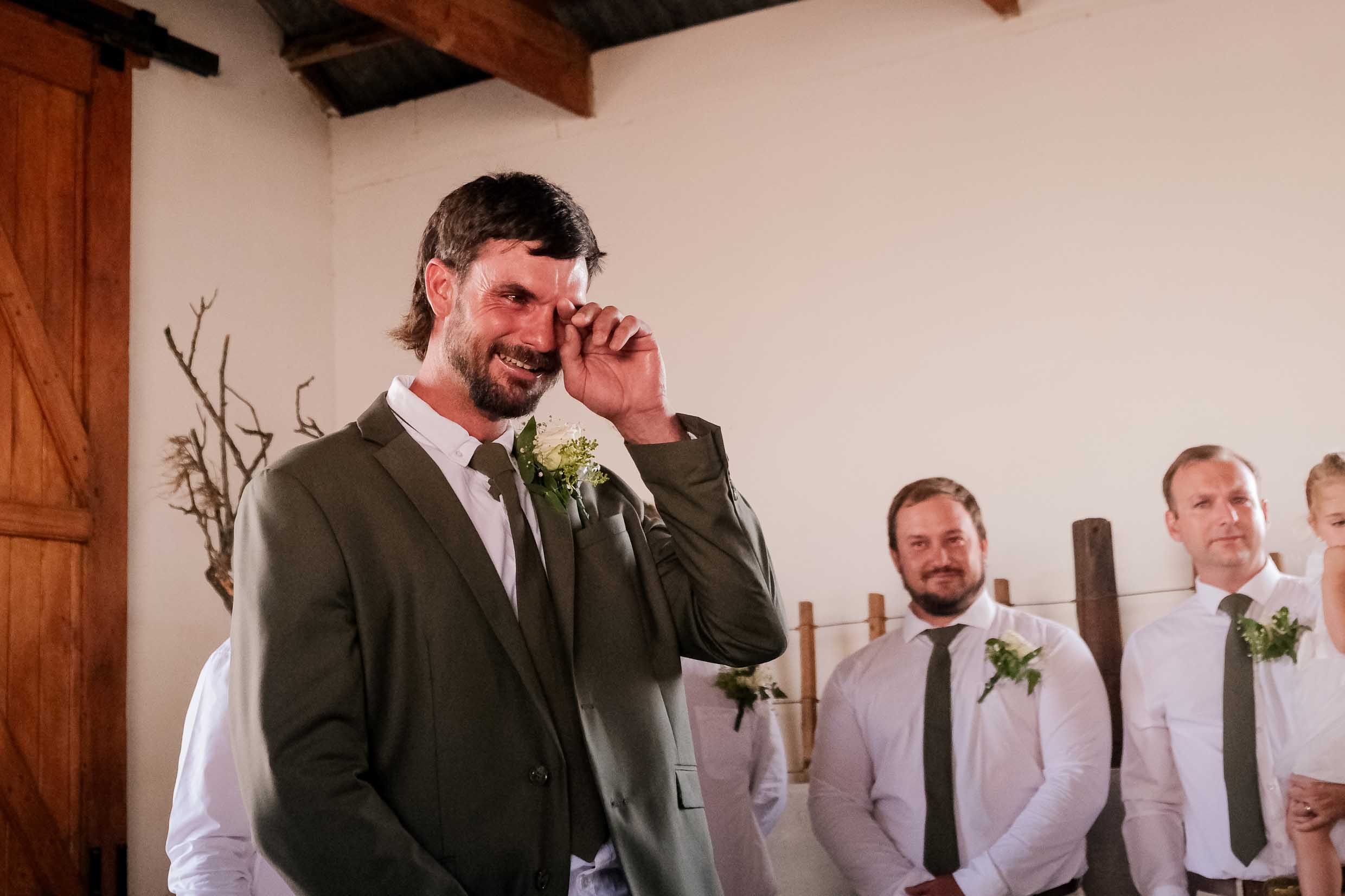 Groom in an olive green suit wiping away happy tears as he smiles at the front of a rustic farm ceremony, with his groomsmen looking on, wedding photography by Adri Krige, Eastern Cape wedding photographer